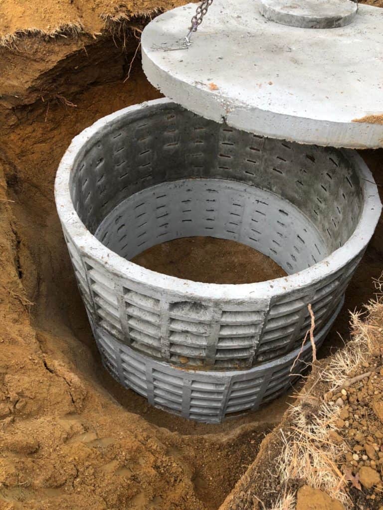 A round concrete well ring, partially buried in the earth, is shown with its concrete cover being lifted—common in NY during cesspool service Suffolk County or septic system services Suffolk County.