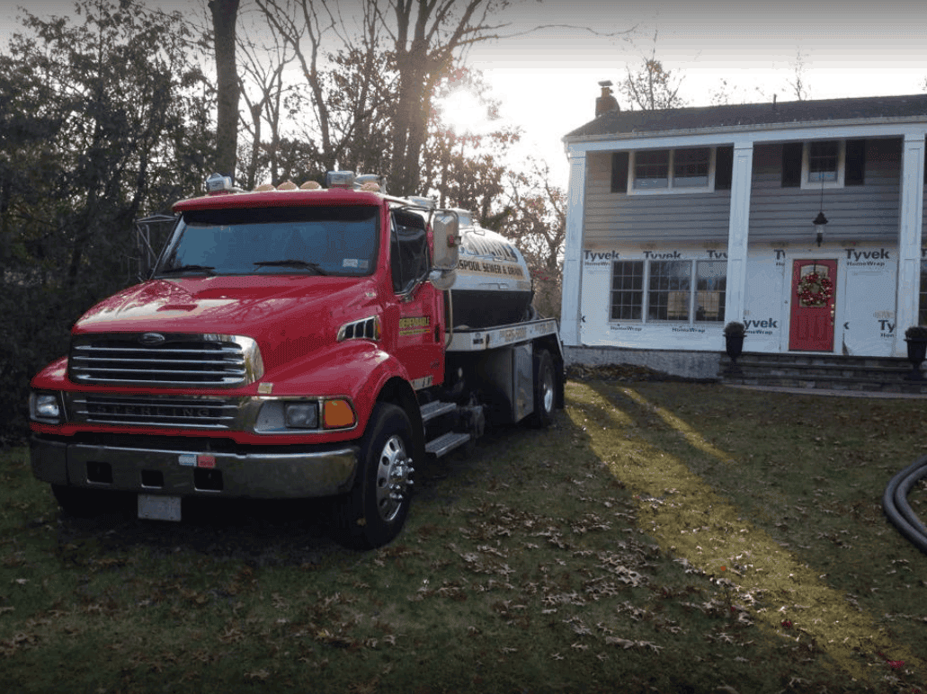 A red water delivery truck is parked on the lawn in front of a two-story NY house with Tyvek wrap, indicating construction or renovation work. Trees and scattered leaves are visible around the yard.