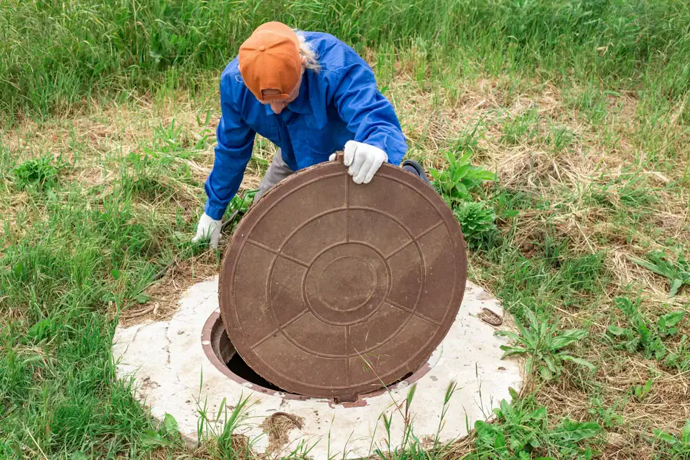 A person in a blue shirt, orange cap, and white gloves lifts a round manhole cover in a grassy outdoor area, revealing an open hole beneath.