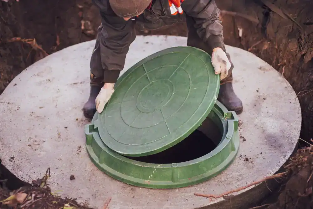A person wearing gloves and work clothes is lifting the green lid of a concrete septic tank or underground utility access point.