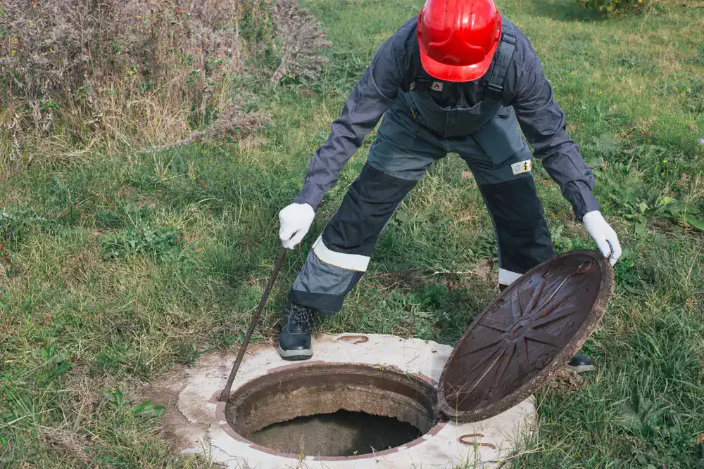 A worker in protective clothing and a red hard hat lifts the lid off a manhole in a grassy area using a metal tool.
