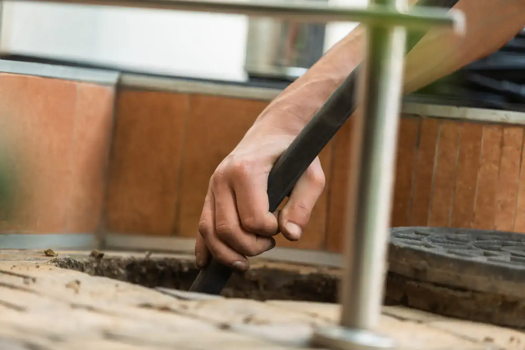 A close-up of a person’s hand holding a black hose or pipe, inserting it into an open drain or manhole, with tiled walls and a metal cover nearby.