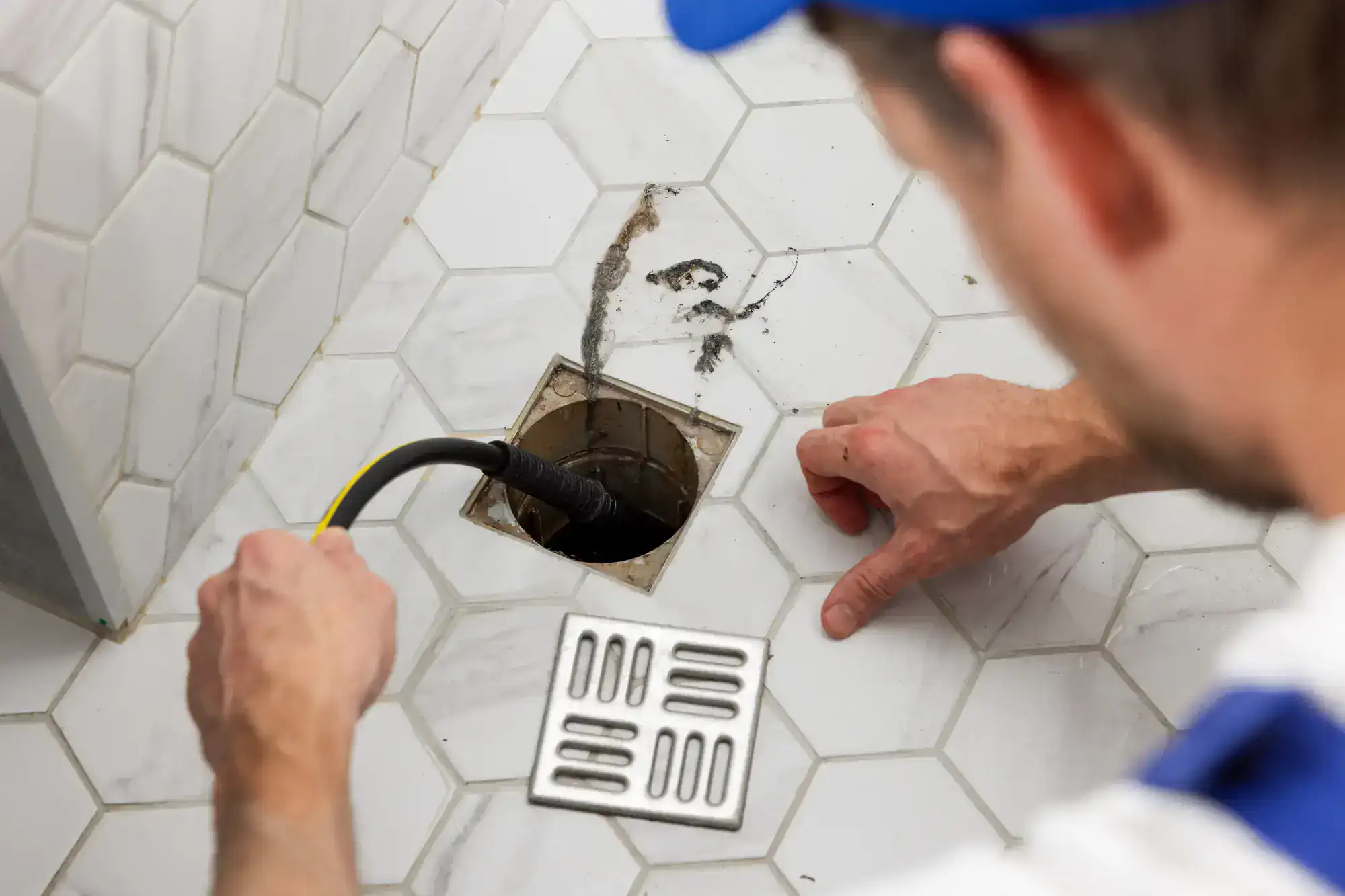 A person uses a drain snake to clear a clogged floor drain in a tiled bathroom. A metal grate and some debris are visible near the drain opening.