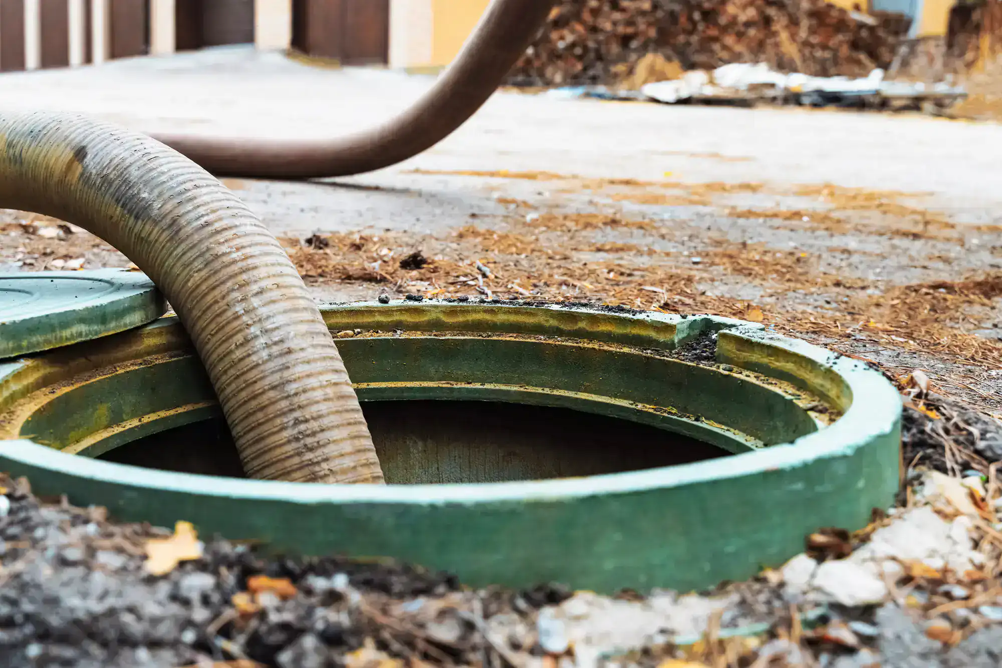 A large hose is inserted into an open green manhole cover outdoors, likely for septic tank cleaning or sewage maintenance. The surrounding ground is muddy with scattered debris.