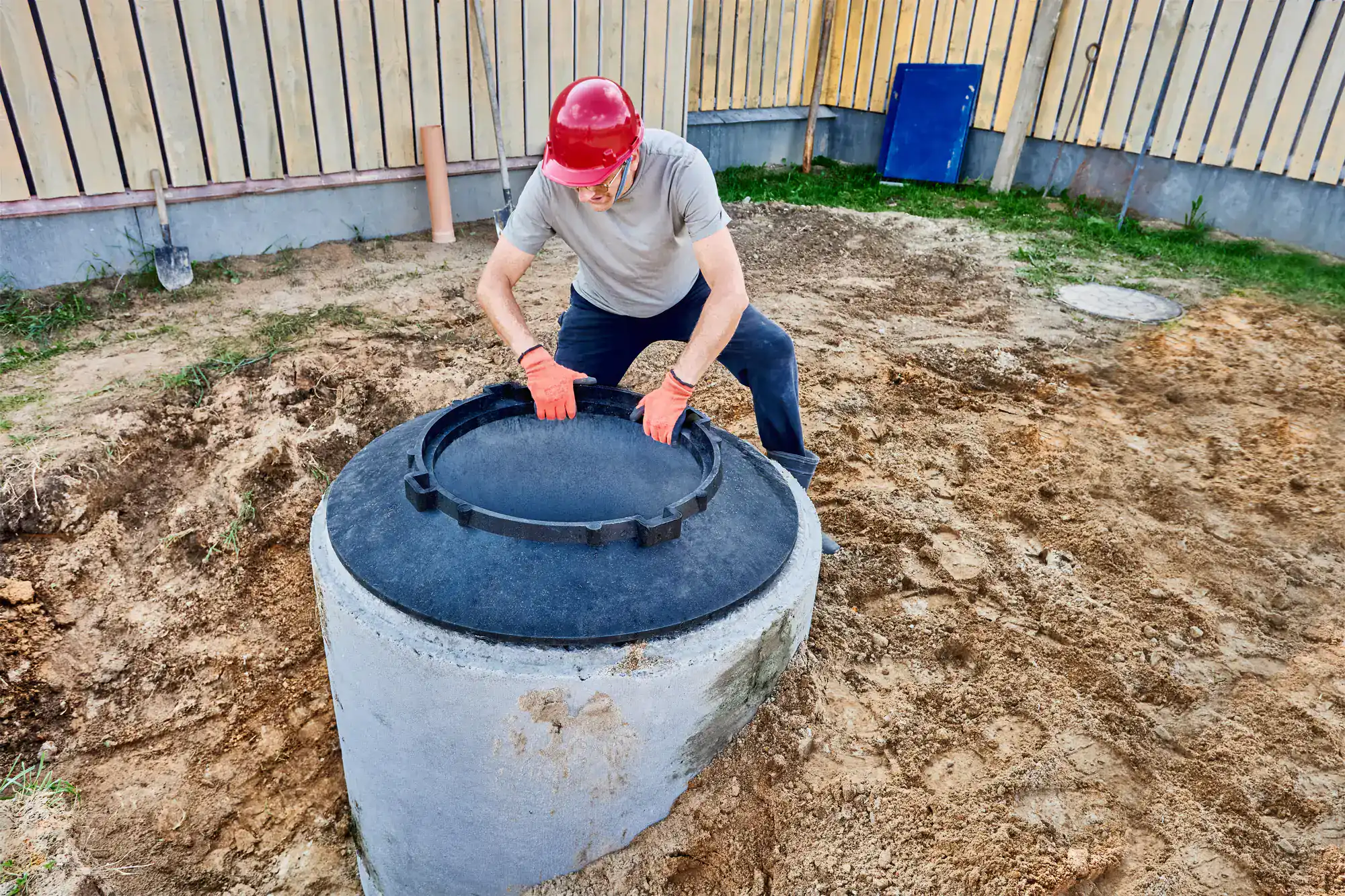 A person wearing a red hard hat and orange gloves is lifting the black lid of a concrete well or septic tank in a backyard, possibly performing cesspool service in Suffolk County, NY. A wooden fence and some dirt surround the area.