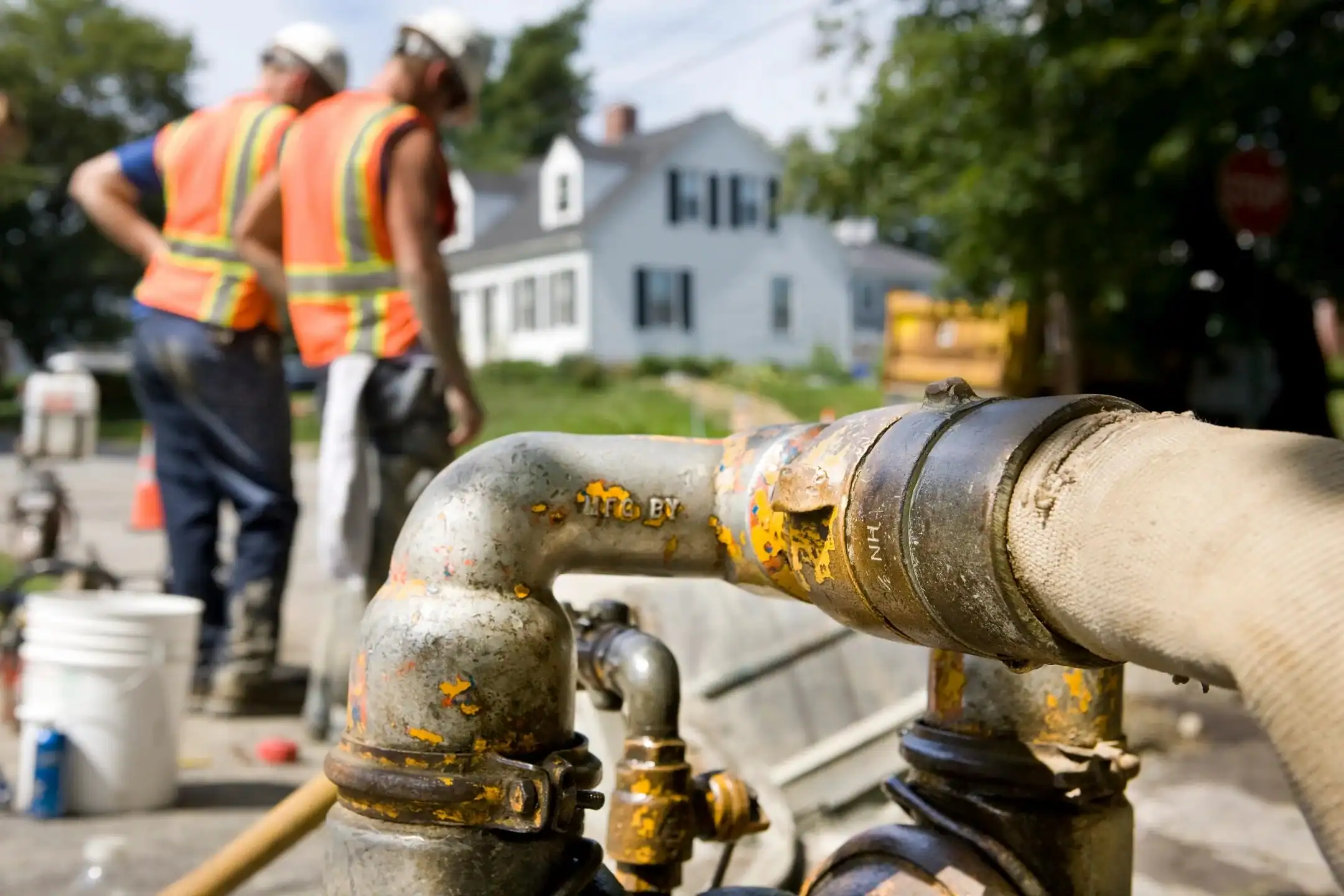 Close-up of a metal pipe with yellow paint and an attached hose, with two construction workers in orange safety vests and hard hats blurred in the background on a residential street.