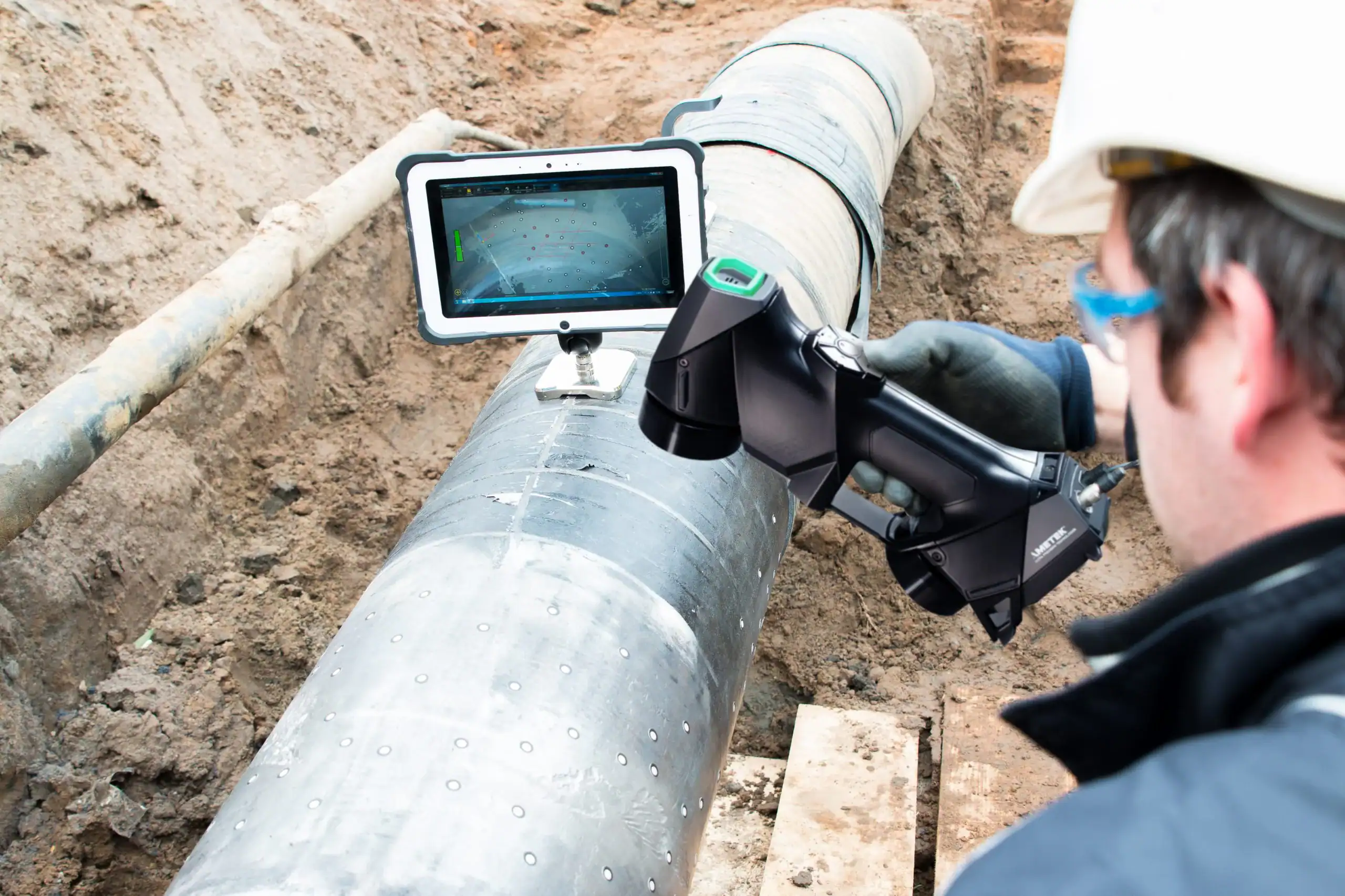 A person in safety gear uses a handheld inspection device and a tablet to examine a large metal pipeline exposed in a trench at a construction site.