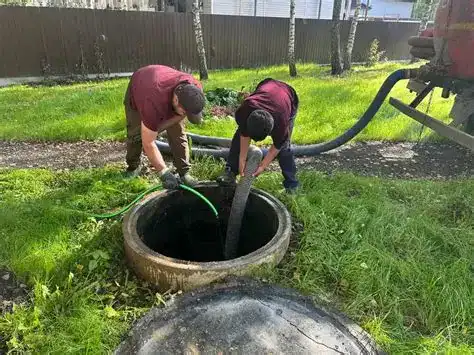 Two workers in maroon shirts are inserting large hoses into an open outdoor sewer or septic tank, surrounded by grass, for maintenance or cleaning.
