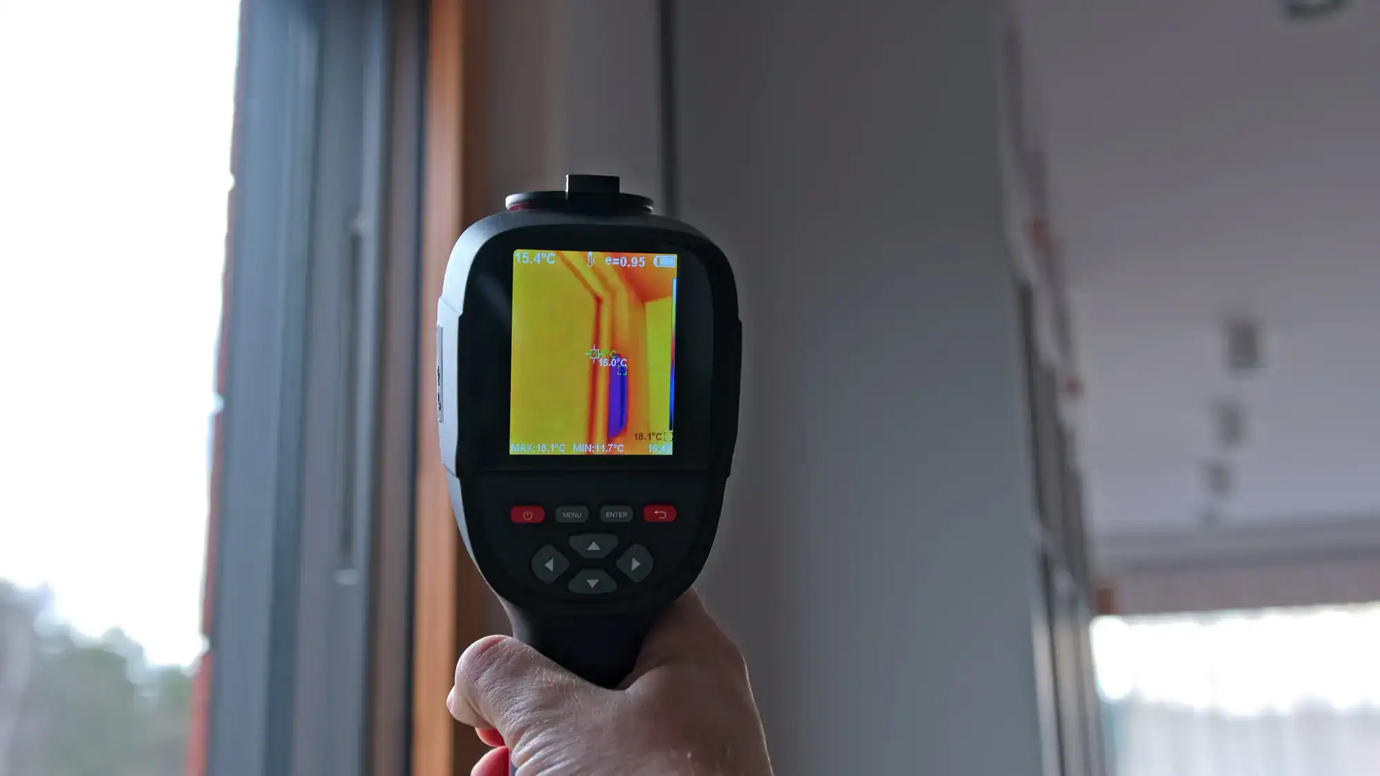 A person holds a thermal imaging camera aimed at a window, displaying temperature differences on its screen, with warmer and cooler areas highlighted in various colors.