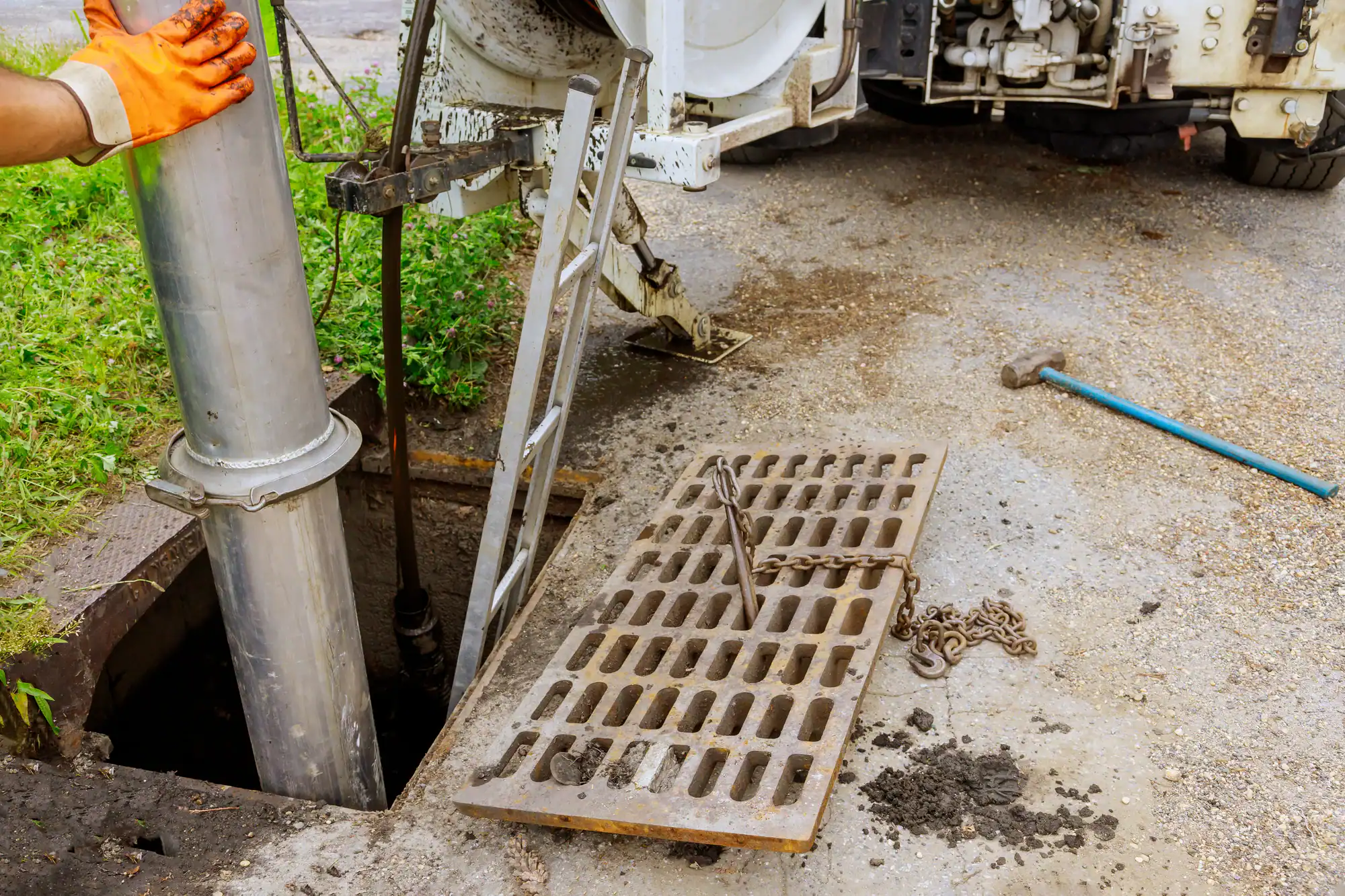 A worker’s gloved hand holds a large vacuum pipe above an open storm drain. Nearby are a ladder, a metal grate with a chain, and a blue crowbar, with a utility truck parked in the background.