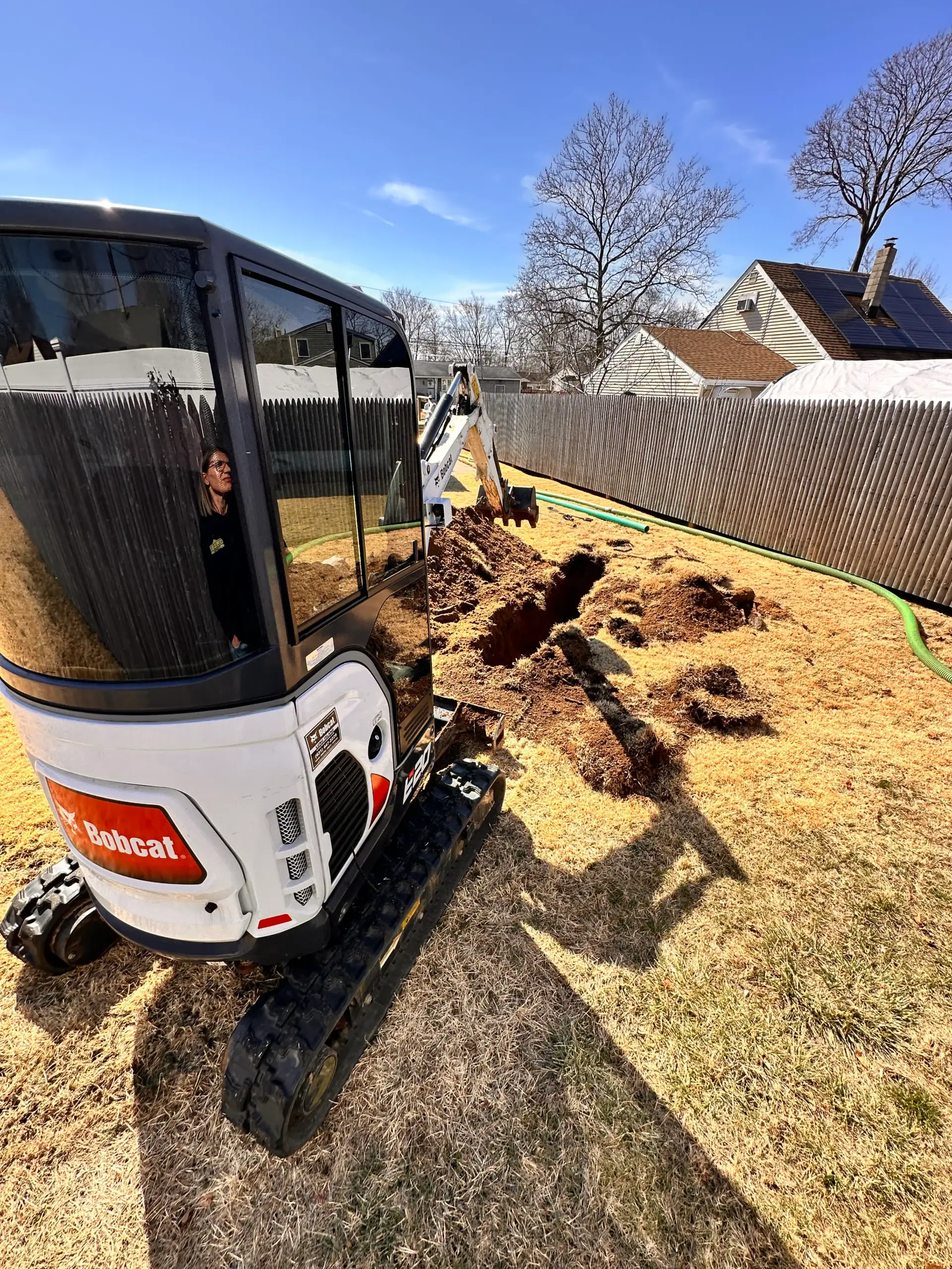 A person operates a small Bobcat excavator in a backyard, digging a long trench in the brown grass near a wooden fence, with houses and leafless trees in the background under a clear blue sky.