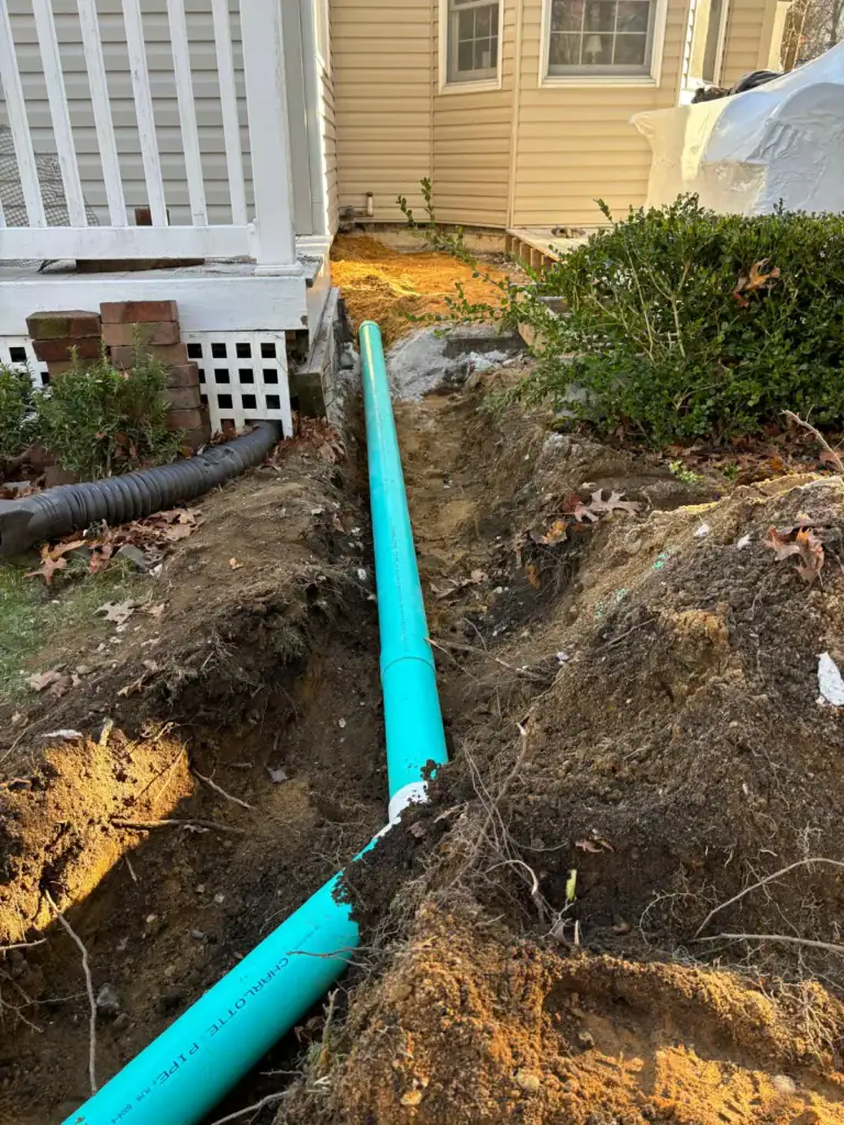 A large turquoise pipe is being installed in a trench beside a house with tan siding and white trim. The trench runs alongside the porch and landscaped yard. Dirt and leaves are scattered around the exposed area.