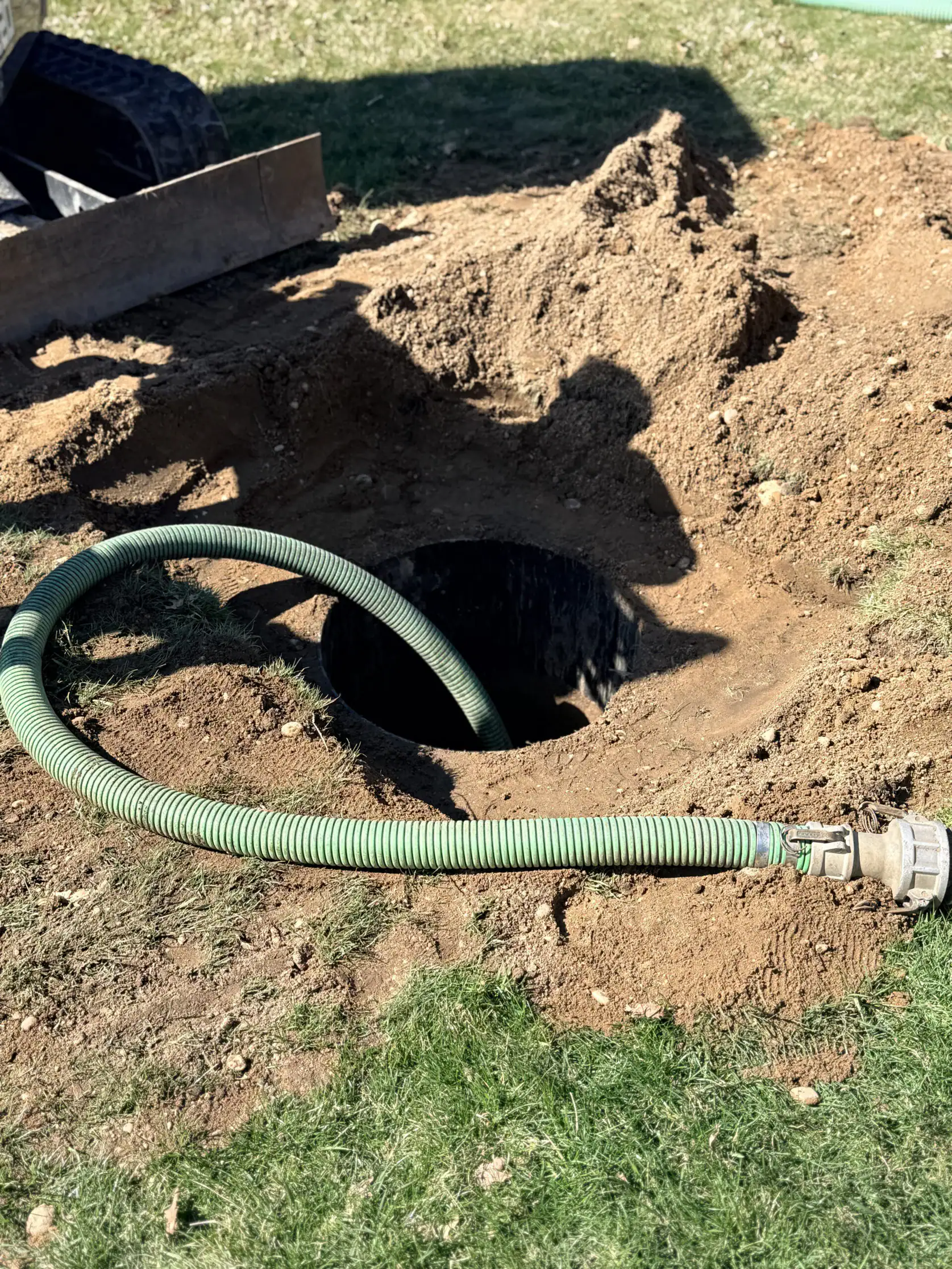 A green hose is inserted into a large hole in the ground, surrounded by loose dirt and grass. The scene appears to be part of some outdoor excavation or maintenance work.