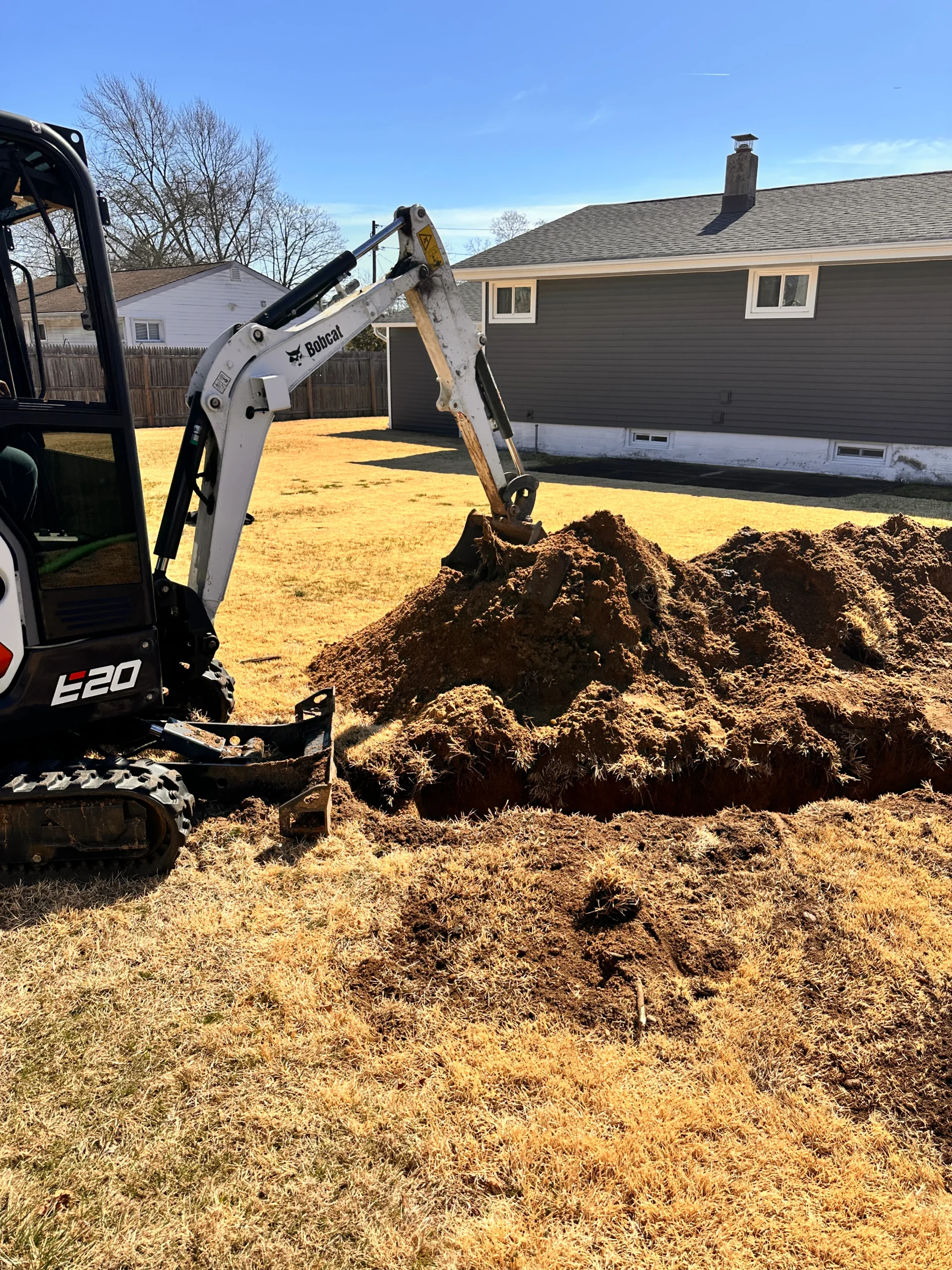 A small excavator is digging a trench in a yellowed grass yard, creating a large pile of dirt near a gray house with white trim in the background on a sunny day.