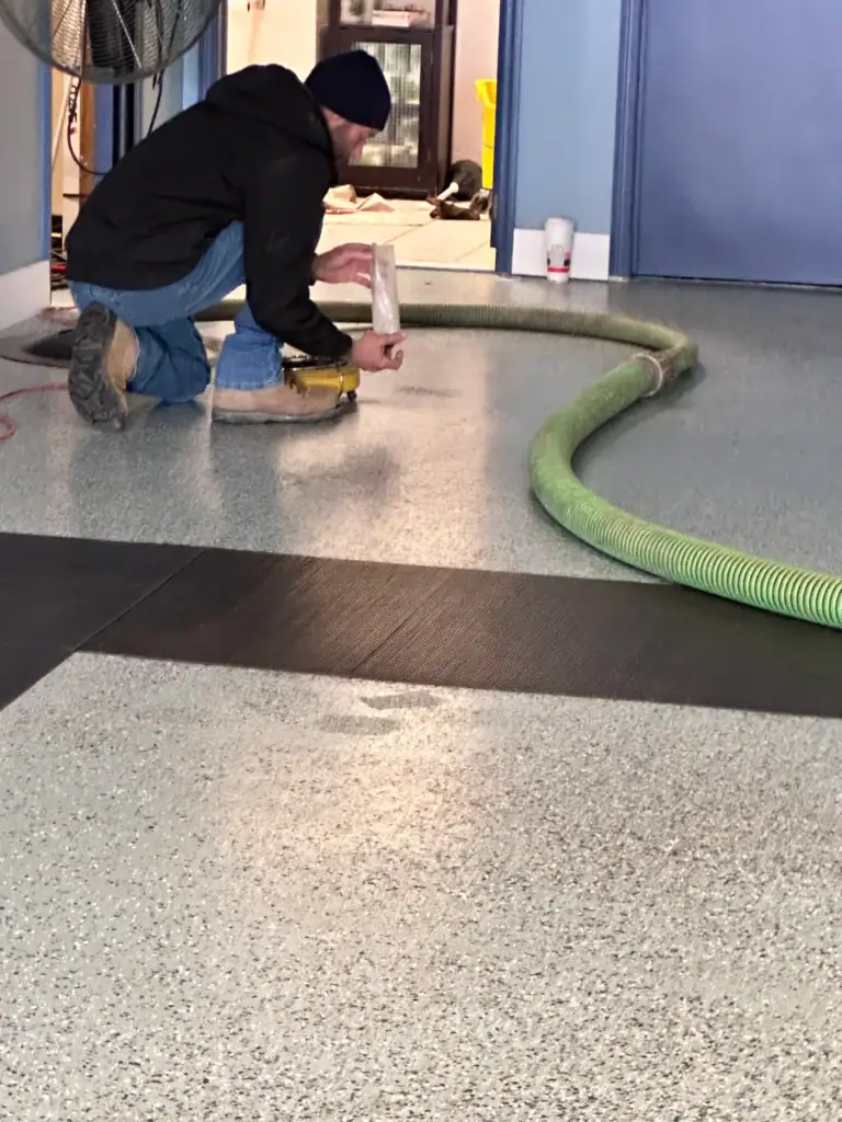 A person wearing a black jacket, jeans, and a beanie is kneeling on a speckled floor, working with a hose and a tool near a blue door. A large green hose runs across the floor.