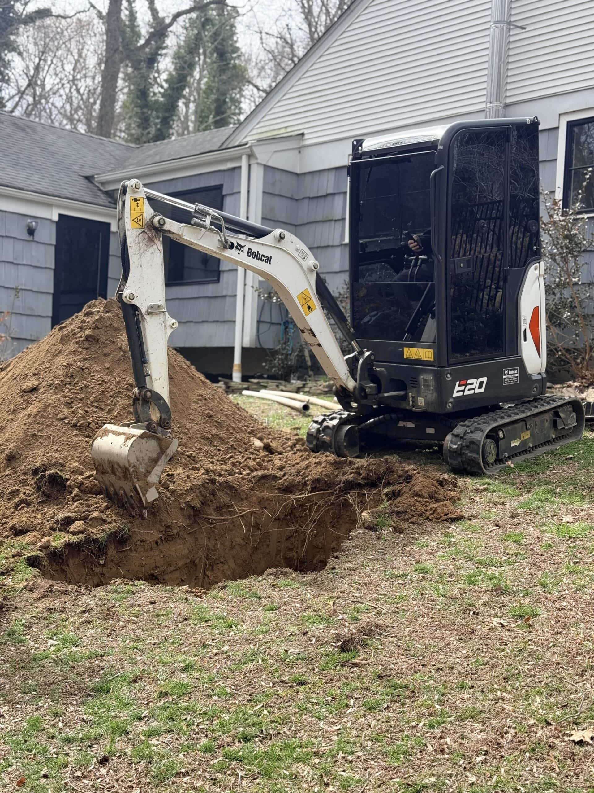 A small excavator is digging a hole next to a house, with a large pile of dirt beside it on the grass. The house has gray siding and white trim. Trees are visible in the background.