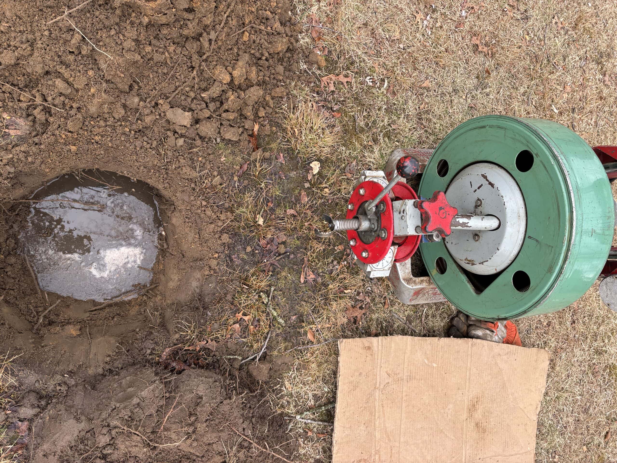 A green and red mechanical tool is positioned on the ground next to a dug hole partially filled with water. The surrounding area has grass, dirt, and a piece of cardboard.
