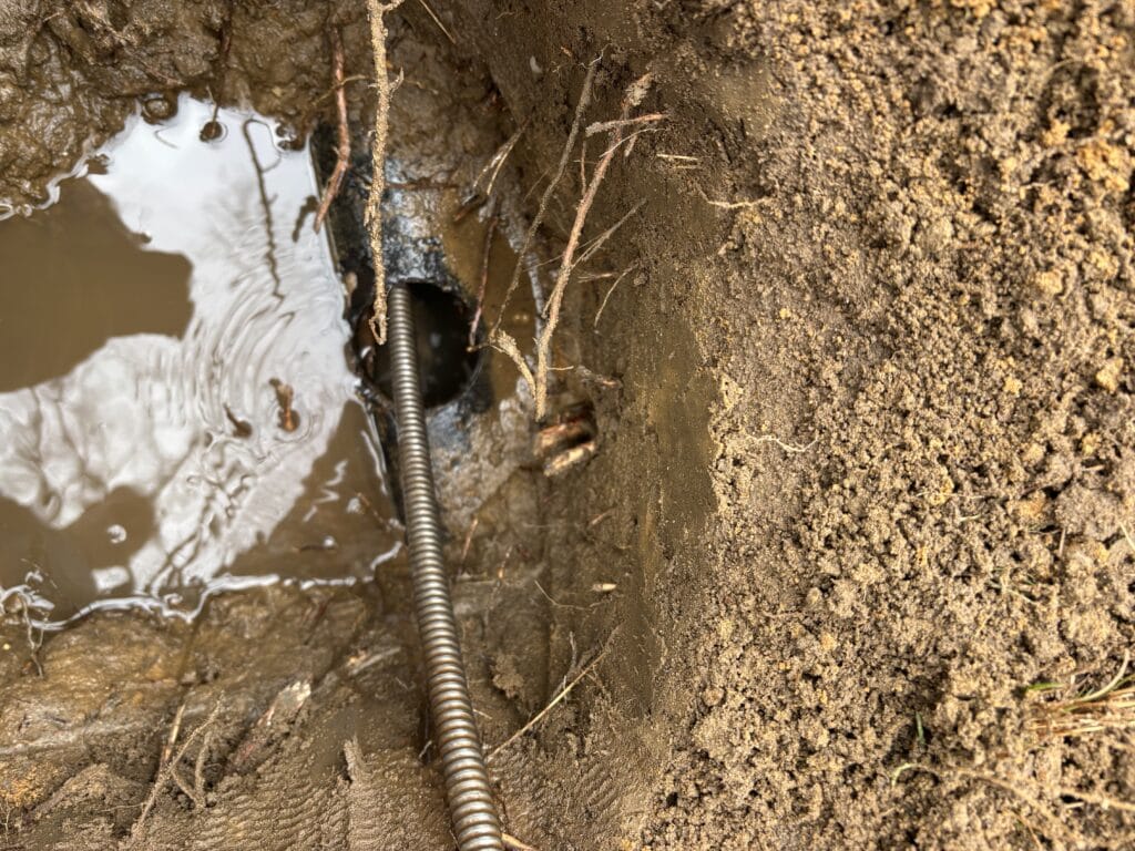 A metal snake tool enters a muddy hole in the ground next to standing water and exposed soil, likely for plumbing or pipe repair work. Small roots are visible around the wet area.