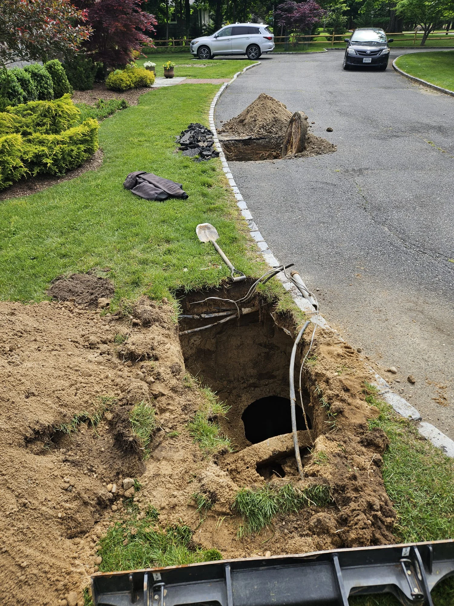 A large hole dug in a grassy area next to a driveway, with a pile of dirt, exposed pipes or cables, a shovel, and tools scattered nearby. There are two piles of dirt further along the driveway.
