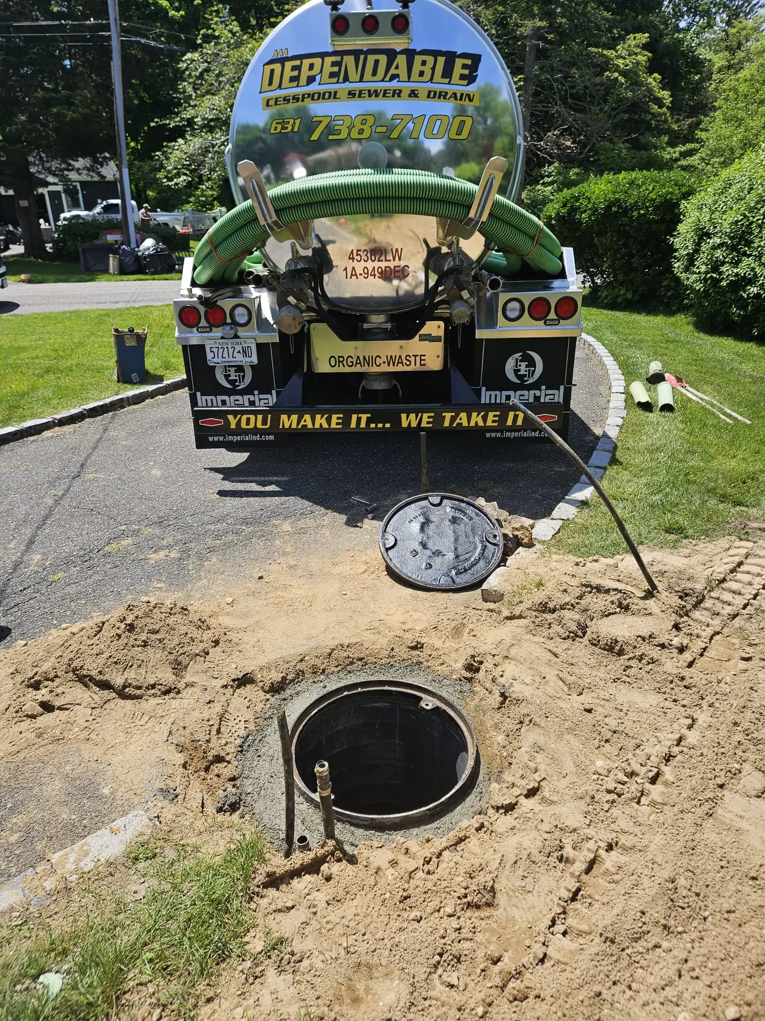 A sewage truck labeled “Dependable Cesspool Sewer & Drain” is parked near an open septic tank on a residential driveway, with tools and sand around the exposed hole.