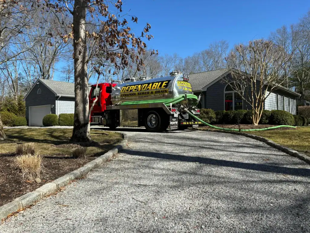 A large "Dependable" service truck is parked on a gravel driveway in front of a gray house, with hoses extended from the truck toward the building. Leafless trees and winter landscaping are visible.