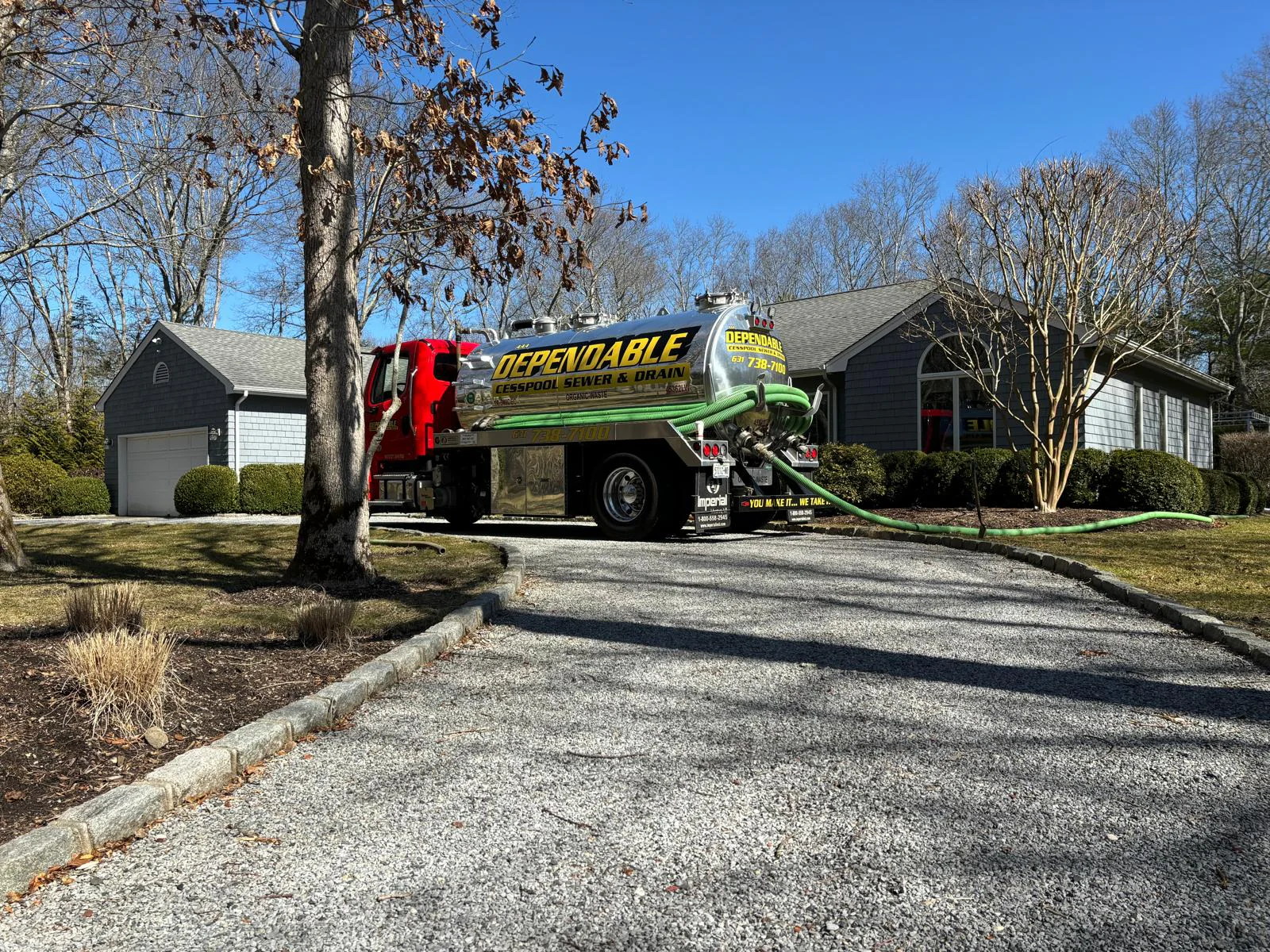 A large "Dependable" service truck is parked on a gravel driveway in front of a gray house, with hoses extended from the truck toward the building. Leafless trees and winter landscaping are visible.