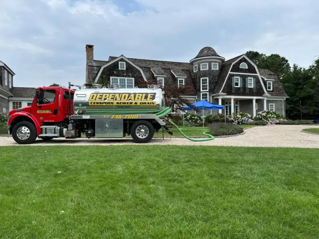 A red septic service truck labeled "Dependable Cesspool Sewer & Drain" is parked on a driveway in front of a large, elegant house with brown siding and multiple gables. A green hose extends from the truck.